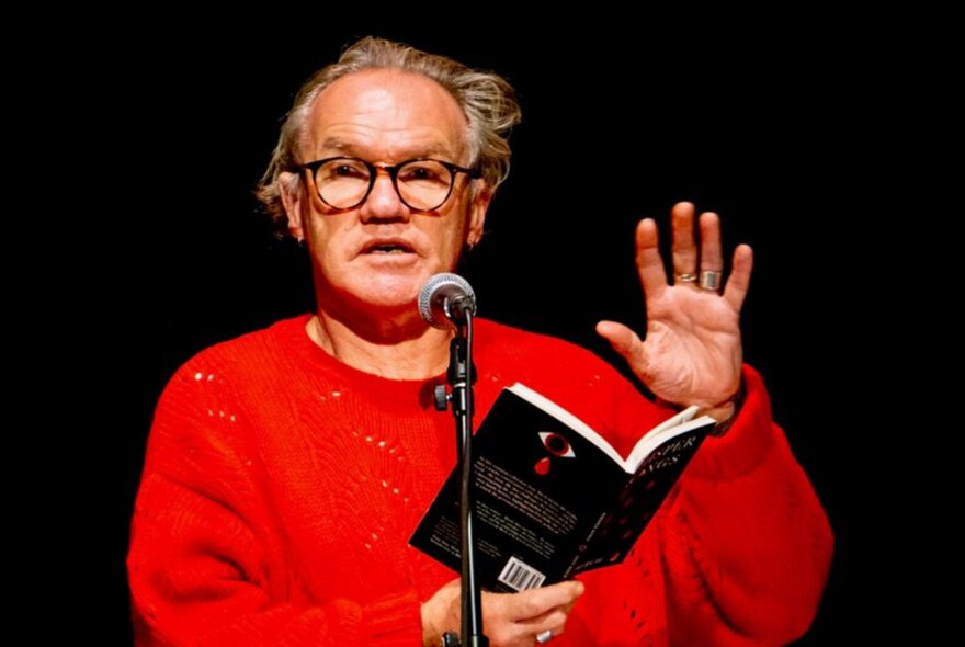 Writer Tony Birch, wearing a red jumper and glasses, reading from a book into a microphone on a stage, in front of a black curtain.