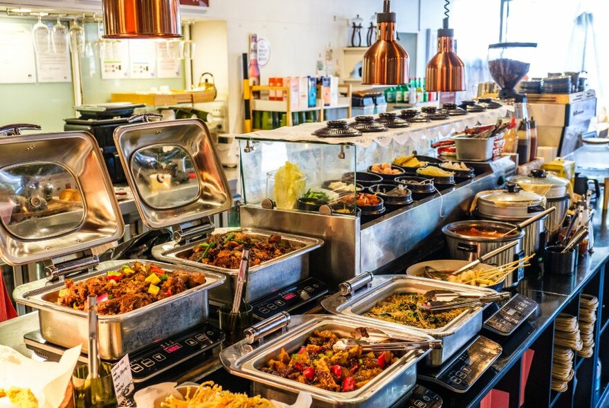 Dishes of hot food in trays on a restaurant kitchen counter, with cooking equipment in the background.