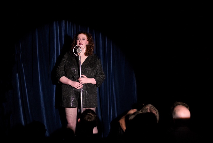 An impro performer on a stage singing into a microphone.