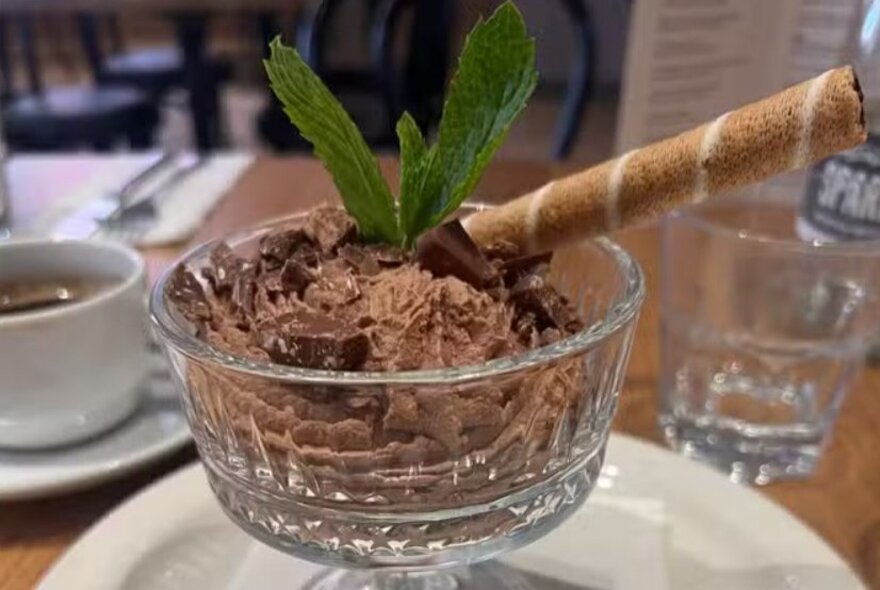 A glass dessert bowl with chocolate mousse, garnished with mint leaves and a poco roll. 