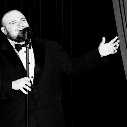 Black and white image of a man in a tuxedo singing into a microphone with one hand extended. 