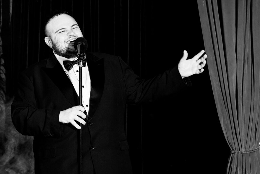 Black and white image of a man in a tuxedo singing into a microphone with one hand extended. 