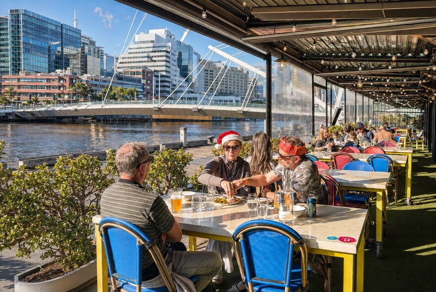 People wearing Christmas hats dining on an outdoor terrace beside the Yarra River with city buildings in the background.