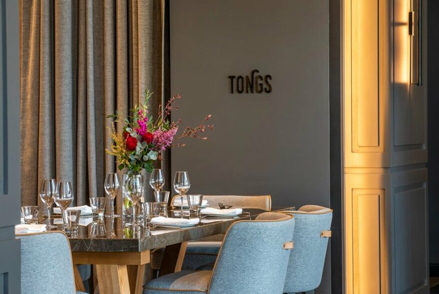 Interior room of a restaurant with a table set for service, light blue upholstered dining chairs, soft curtains and a sign on the wall which reads TONGS.