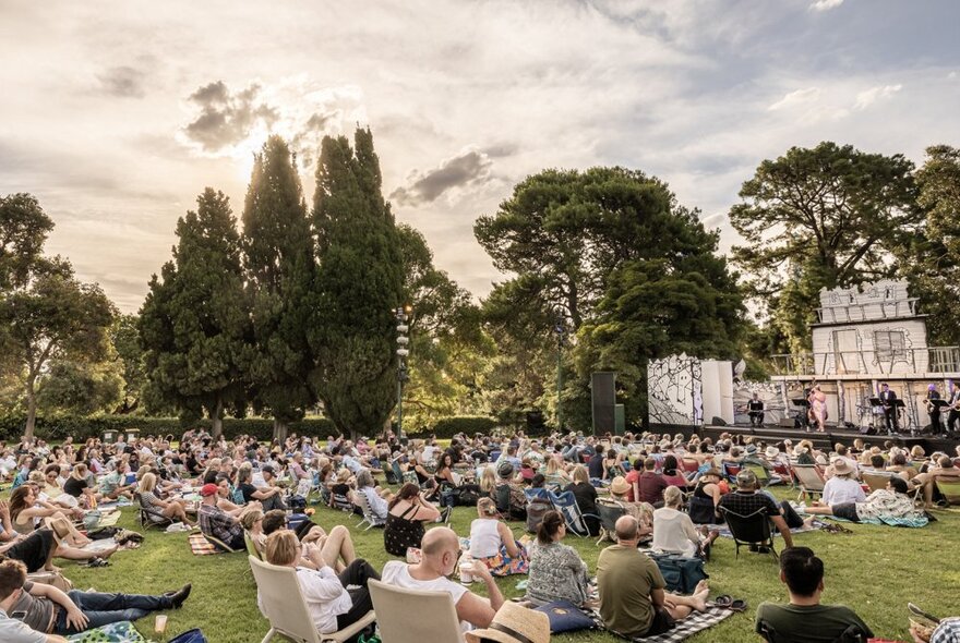 A crowd of people seated on beach chairs and picnic blankets on the lawn at The Royal Botanical Gardens Melbourne, watching performers on a stage.