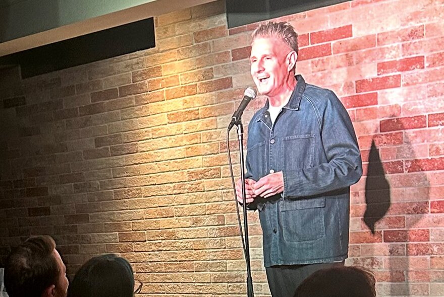 Comedian Wil Anderson performing stand-up at a small venue with a brick wall background. 