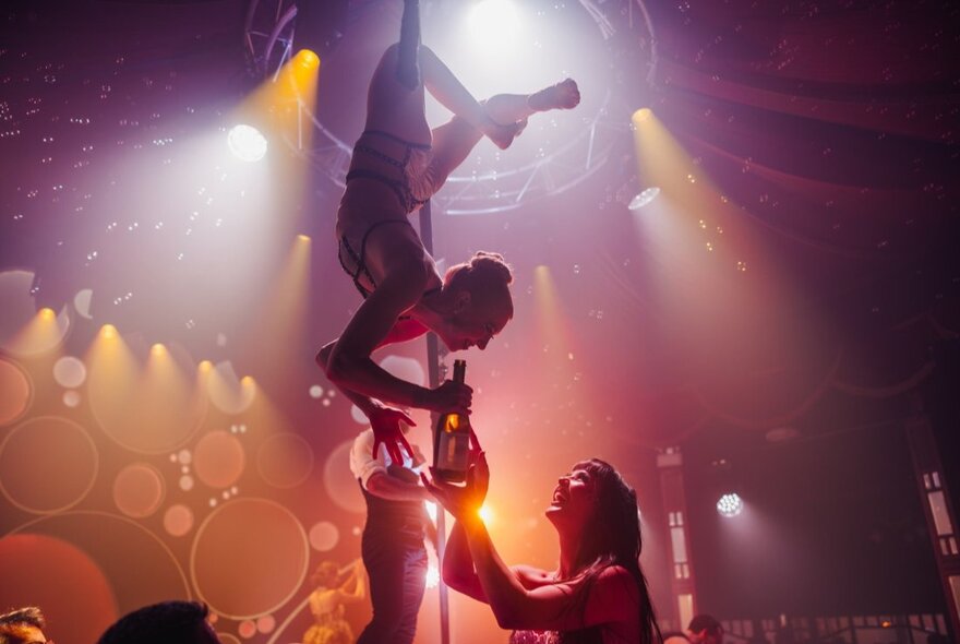 Circus performers on stage in the circular Spiegel Haus, one hanging upside down from a pole as the other passes her a bottle.