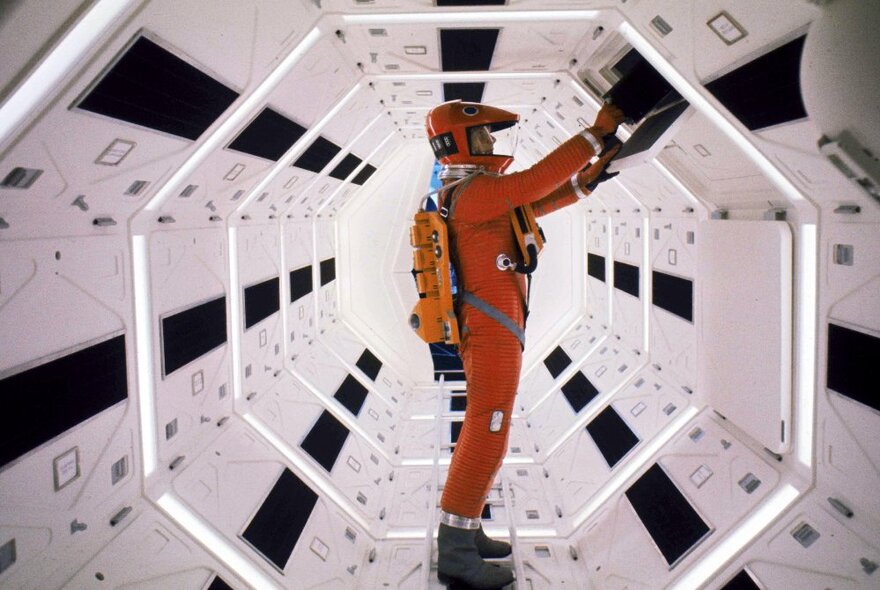 An astronaut in an orange spacesuit, inside a futuristic octagonal white room with white lights and black panels.