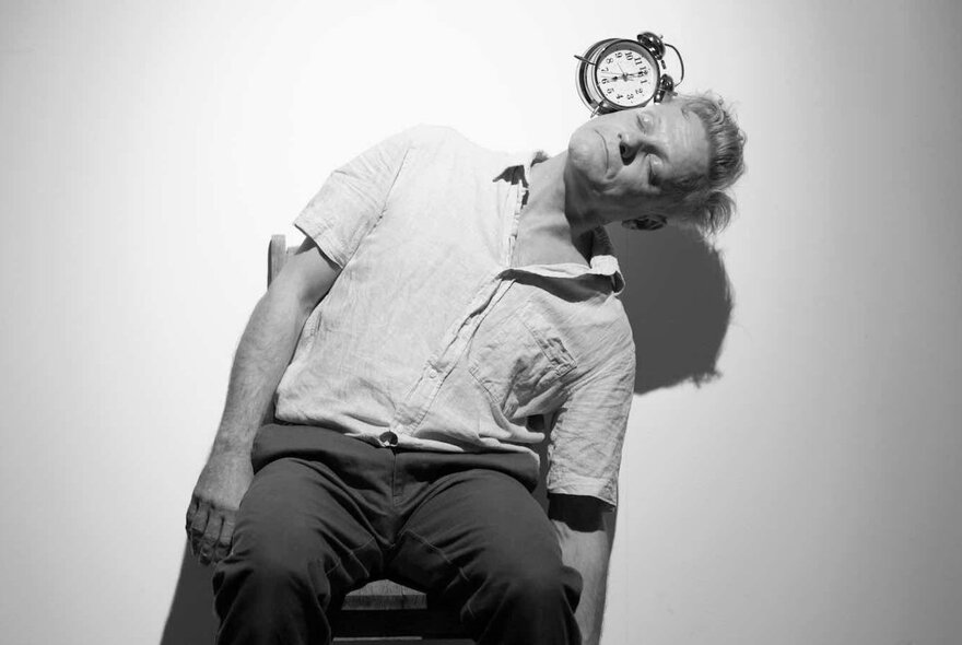 A man sitting on a chair with his back pressed up against a wall, his body leaning to one side, with a small clock resting on the side of his face; a black and white image.