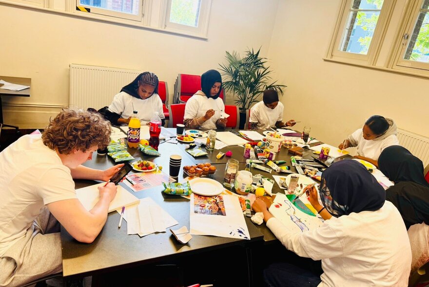 Young adults inside a studio space, creating artworks on canvas, seated around a table with paints and brushes.