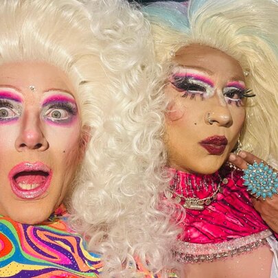 Two drag queens with big blonde hair and pink toned outfits, one looking blankly, the other surprised. 