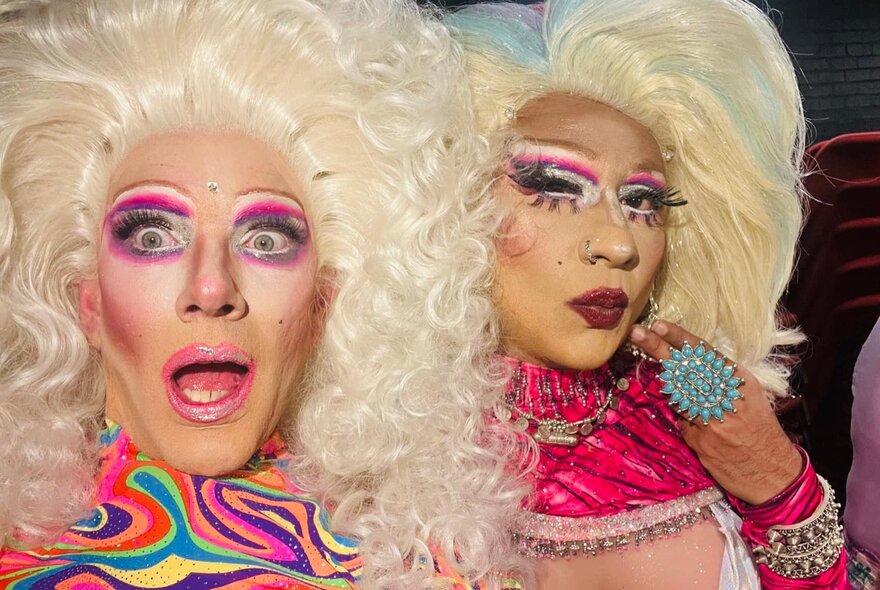 Two drag queens with big blonde hair and pink toned outfits, one looking blankly, the other surprised. 