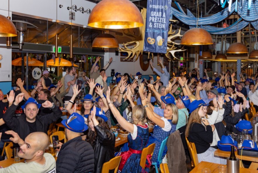 A packed crowd of people cheering with their hands raised inside a lively Bavarian-style beer hall during Oktoberfest celebratons.