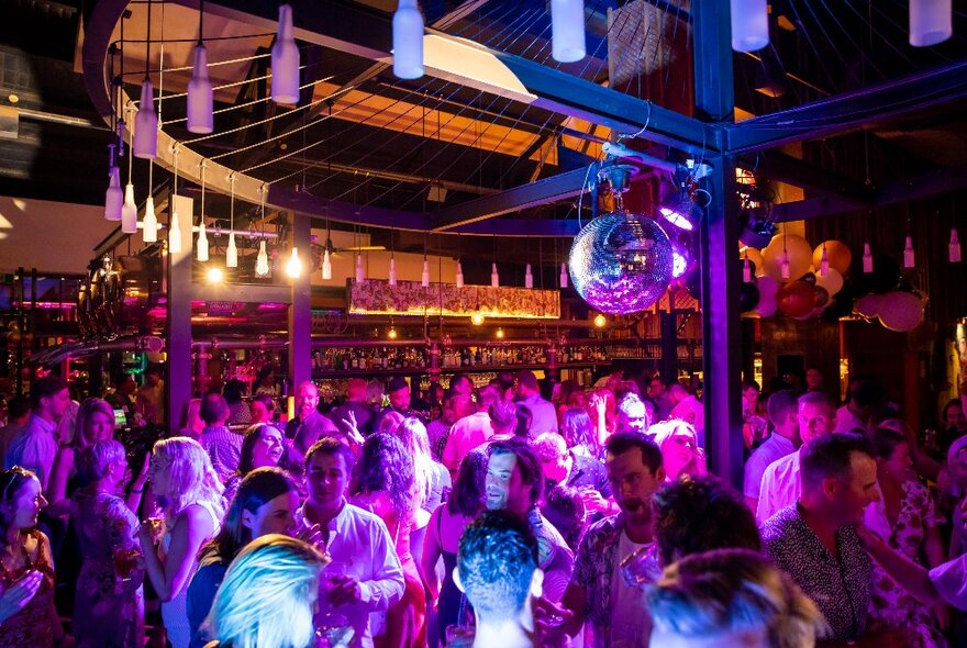 Crowd lit by purple light under mirror balls and lights in a beer garden at night.