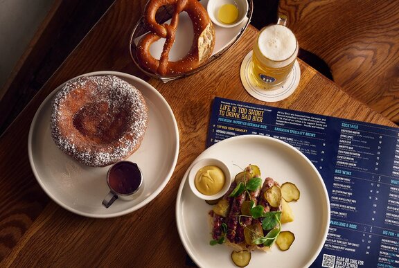 A restaurant table set with bratwurst, pretzel a beer and dessert.