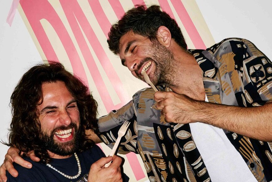 Two Italian chefs laughing with forks in their hands, both with dark hair, one with beard, the other unshaven.