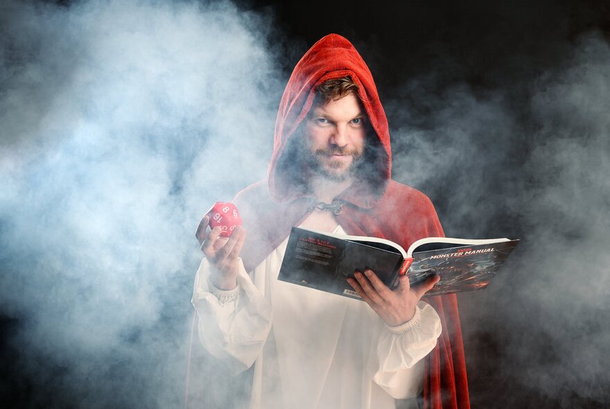 Man wearing a red hood, holding an open book and dice in a misty environment.