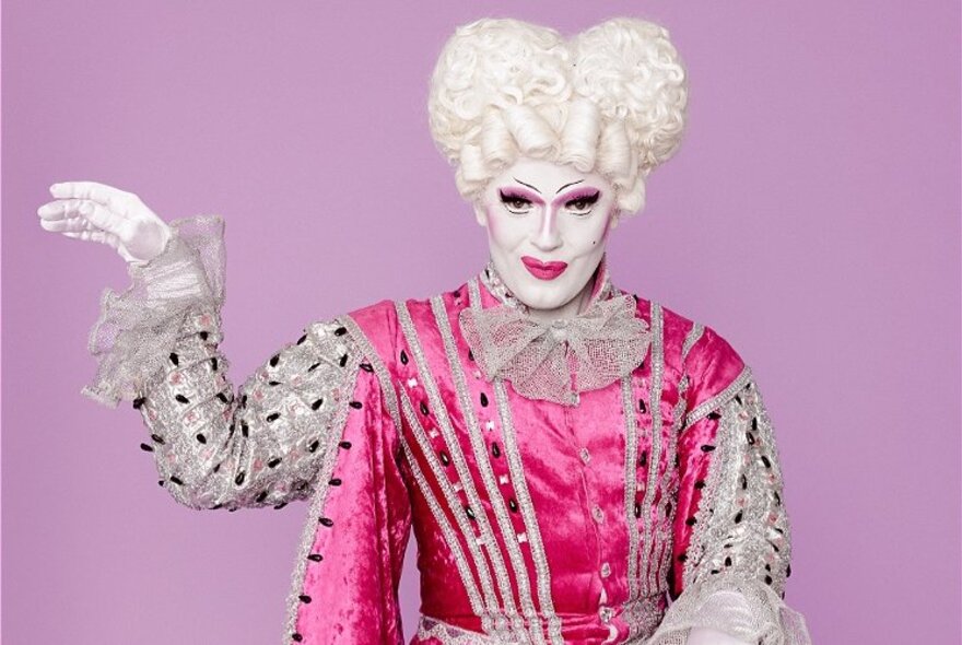 Camp performer wearing white wig and makeup with pink courtier's outfit, with hand raised.