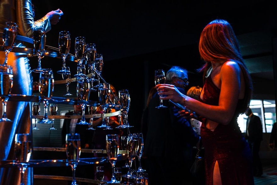 An indoor evening social event with a multi-tiered champagne or sparkling wine glass display set up in dim lighting.