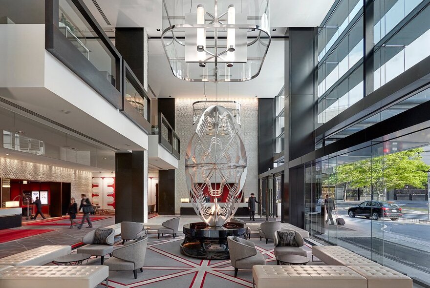 Multilevel hotel foyer with oversized lighting and sculpture, seat and table seating with view of reception desk and trees outside. 