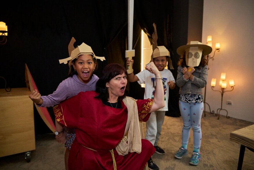 Children and an adult engaging in an interactive gladiator-themed workshop, wearing cardboard helmets and swords.