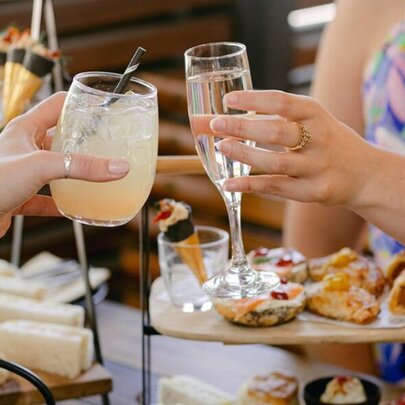 Two hands bringing two drinks together in a clinking motion, with a high tea spread of sweet and savoury food on the table behind them.
