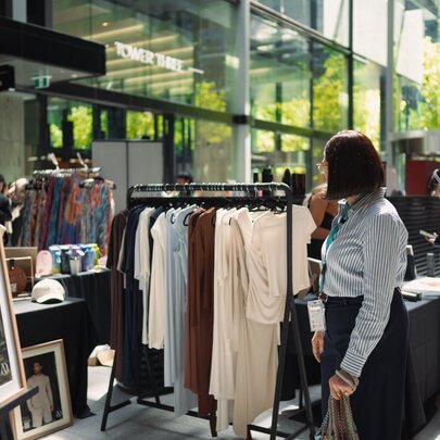 An outdoor fashion and art market event featuring clothing racks, framed artwork, and shoppers browsing stalls.