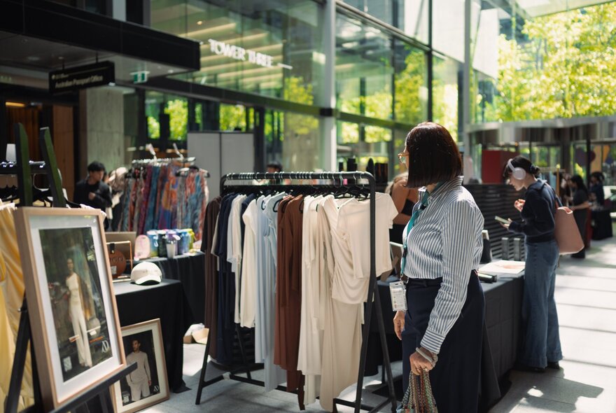 An outdoor fashion and art market event featuring clothing racks, framed artwork, and shoppers browsing stalls.