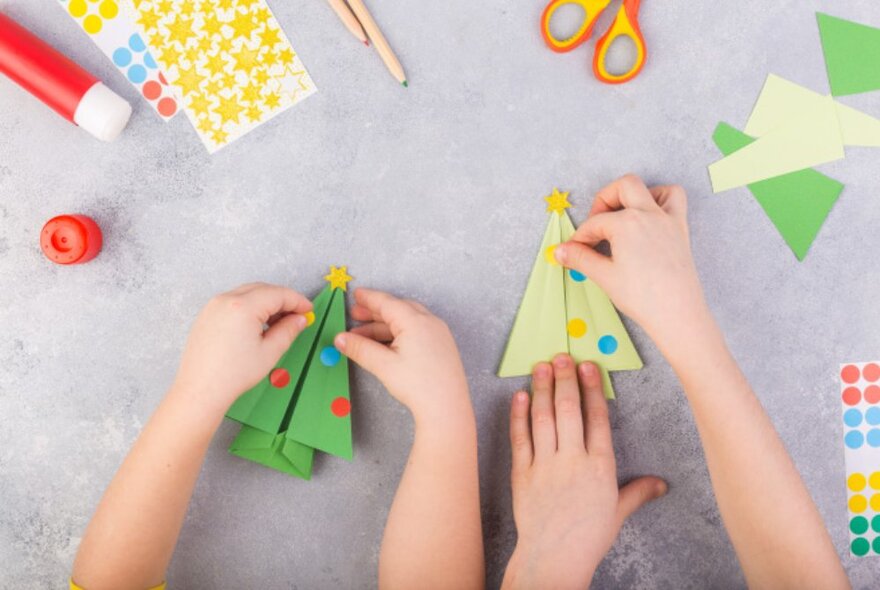 Children's hands folding paper to make Christmas tree decorations with craft materials.