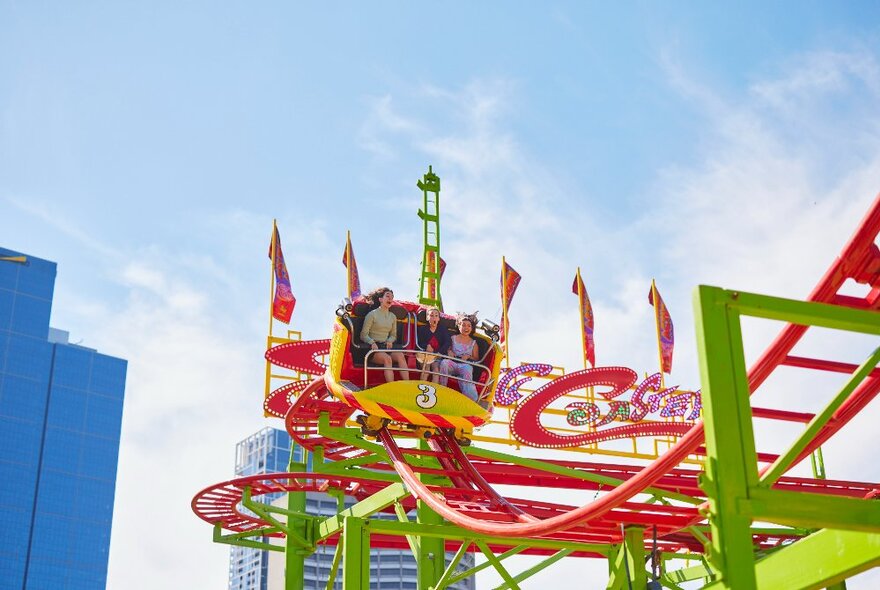 An open air carriage on a roller coaster ride with three people sitting inside the carriage, on a brightly coloured and curved roller coaster track.