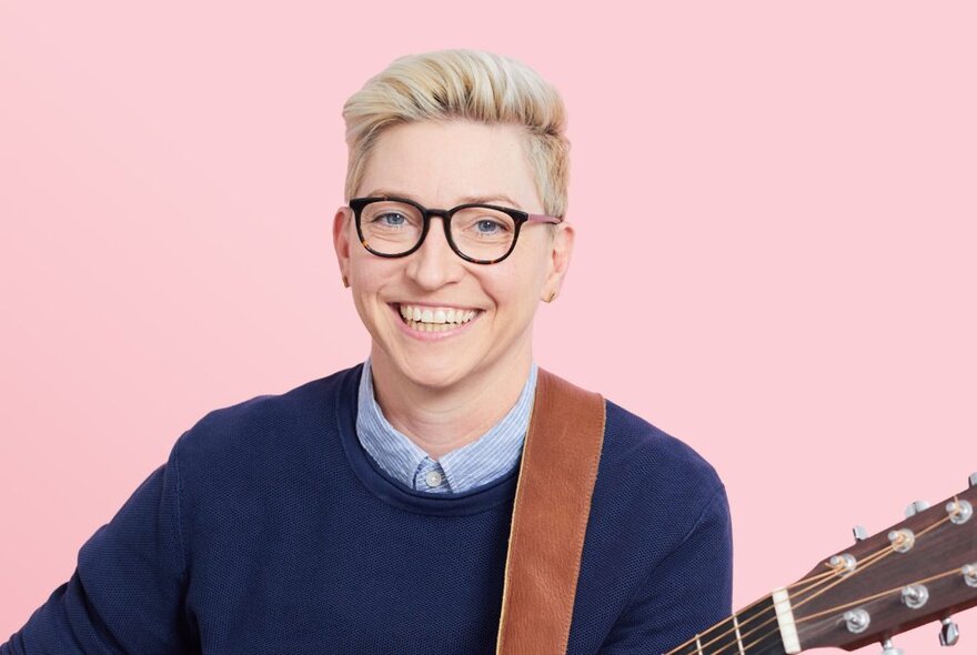 A person smiling at the viewer with short blond hair, wearing glasses and playing an acoustic guitar which is only partly visible.