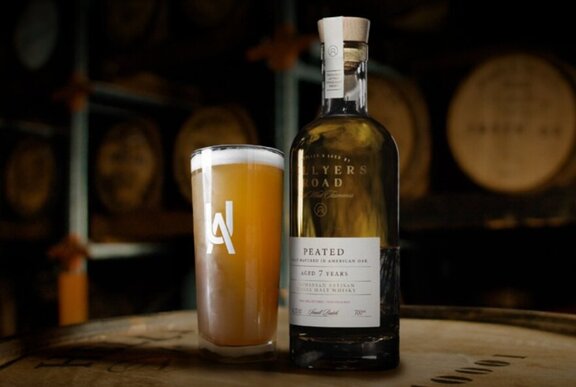 A bottle of whisky and glass of beer on a wooden barrel.