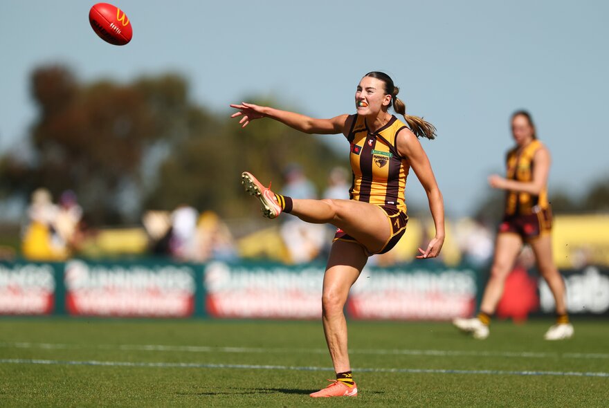 A Hawthorn AFLW player kicking a football with her leg raised high in the air and her arms outstretched.