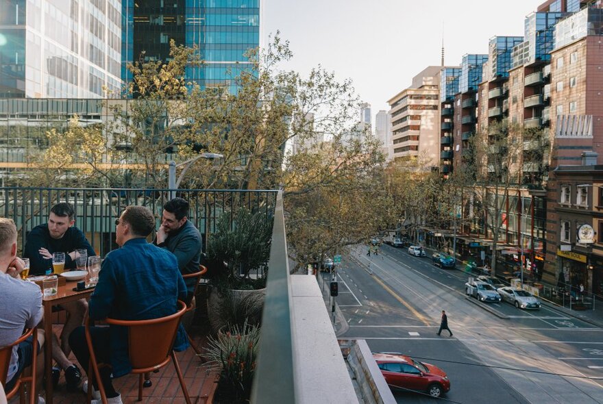 People seated at an outdoor table with drinks on a rooftop overlooking a city street.