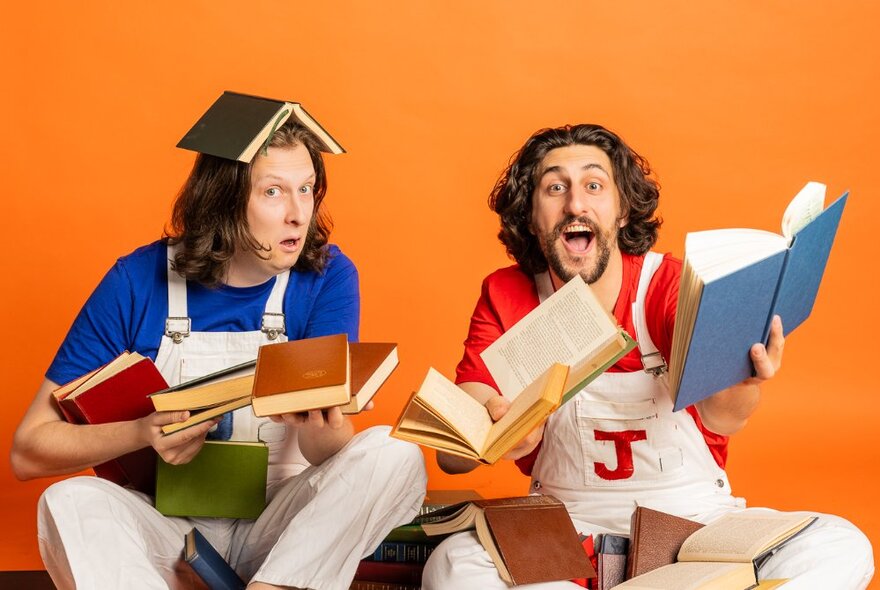 Two comedians in white overalls with bemused and silly expressions on their faces, holding multiple books in their hands, set against an orange background.