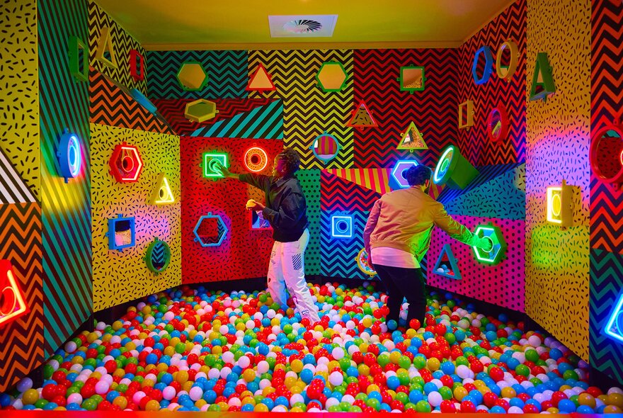 Two people touching colourful shapes on the wall while in a giant ball pit.