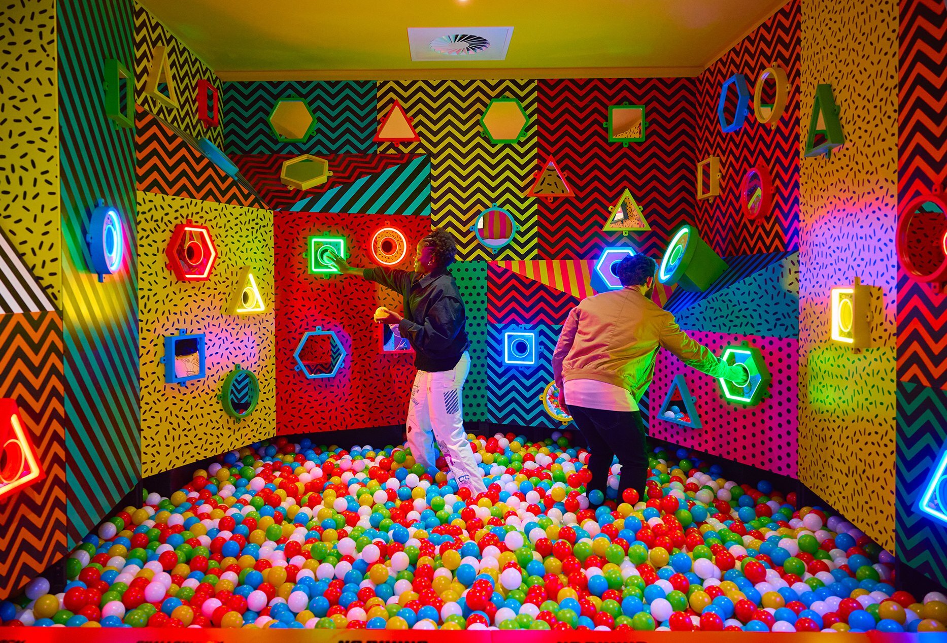 Two people touching colourful shapes on the wall while in a giant ball pit.