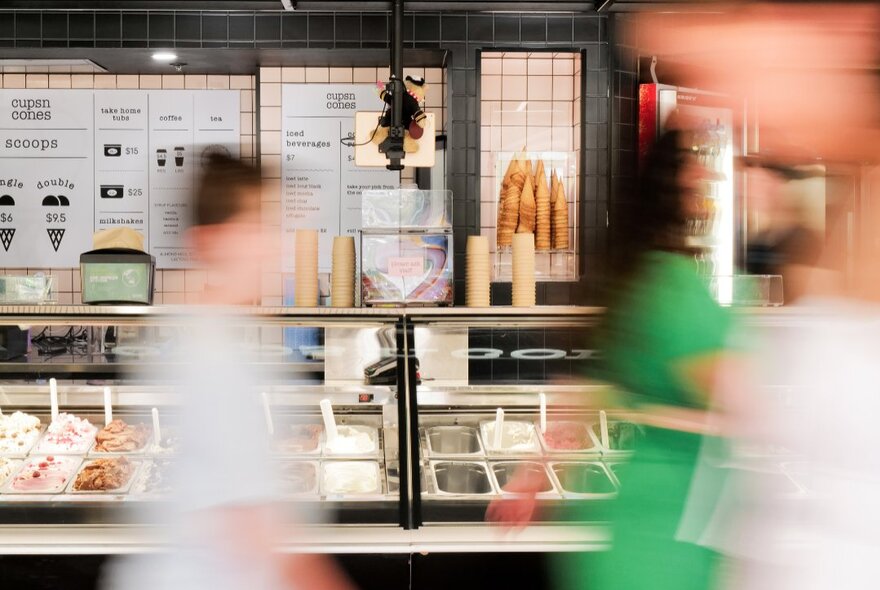 The Cups N Cones kiosk with tubs of gelato in a display fridge, and a menu on the back wall,  with blurred people in motion walking past.