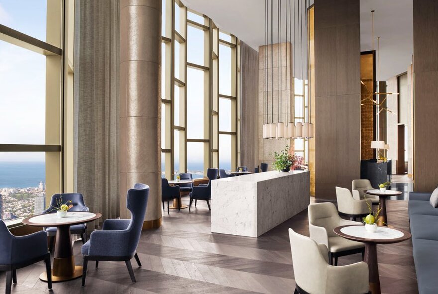The lobby lounge on level 80 of the Ritz-Carlton Hotel, with club armchairs positioned small round tables, muted soft colours and large floor to ceiling windows with views out across the city.