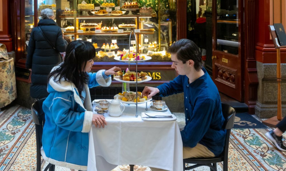 Two people seated at table with white table cloth eating cakes, sandwiches and drinking tea.