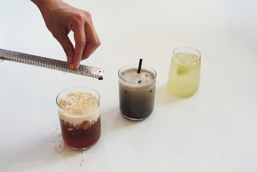 A hand shown using a microplane grater to shave an ingredient over the first of three drinks arranged in a line on a white surface.