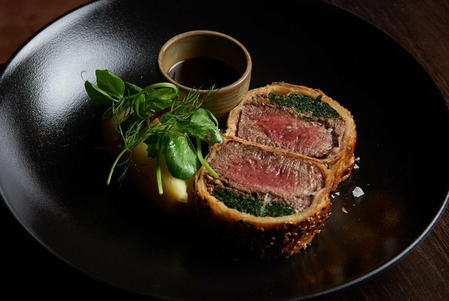 A dark plate with a slice of beef wellington and vegetables.