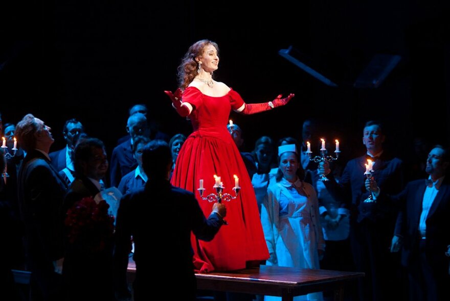 An opera singer performing on stage in a long red dress surrounded by performers out of the spotlight with a blue light cast on them. 