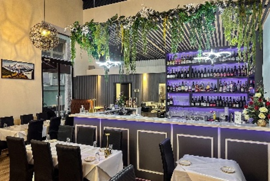 Interior of a restaurant with purple lighting behind the bar and hanging plants; tables set with white tablecloths and black chairs.