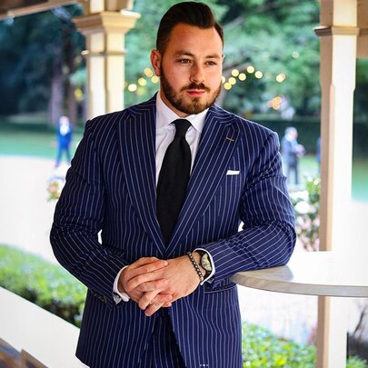 A man leaning on a small table under a verandah wearing a smart blue pinstripe suit.