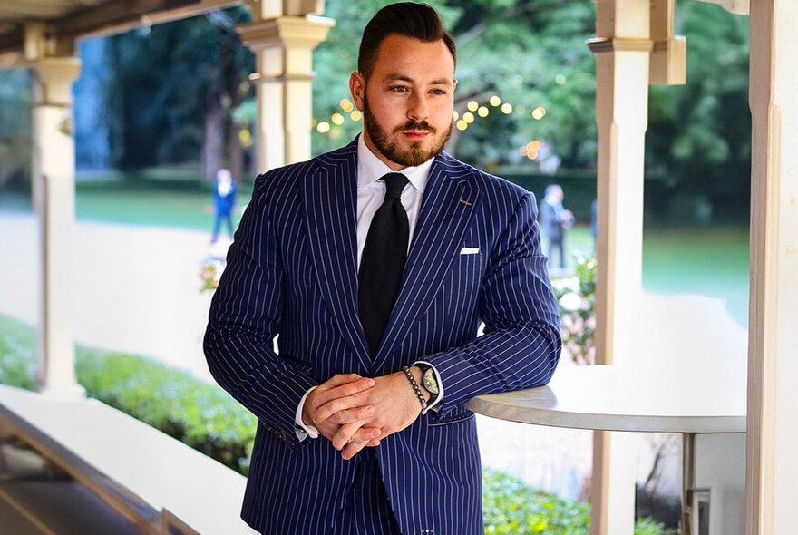 A man leaning on a small table under a verandah wearing a smart blue pinstripe suit.