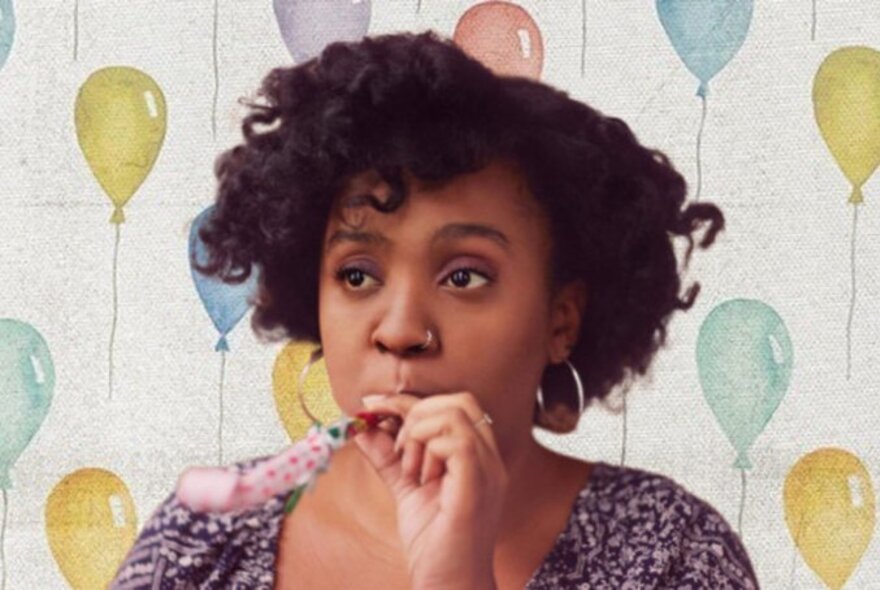 Comedian Chanel Ali, blowing into an inflated and extended party blower that she holds to her lips, with a quizzical expression on her face, a backdrop of painted balloons behind her.