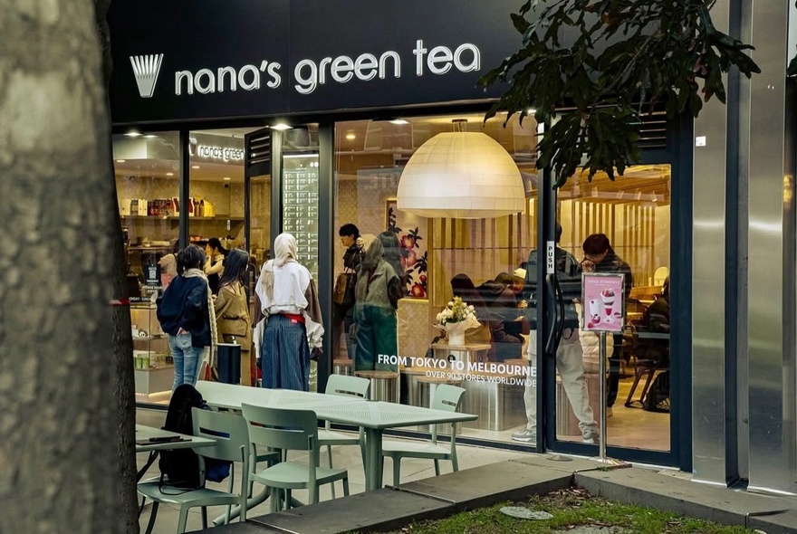 People queuing at the door of a branded cafe with outdoor seating and large glass frontage.
