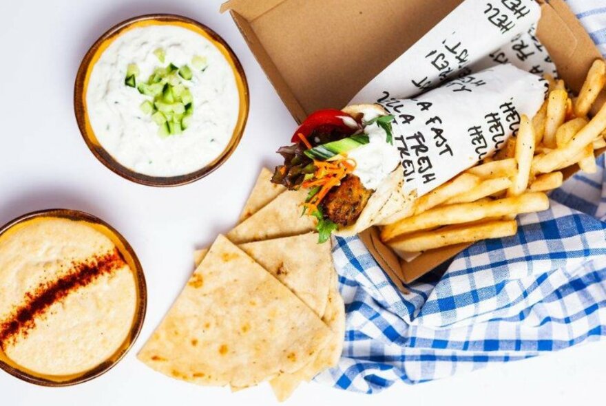 A souvlaki and chips in a box on top of a blue and white checkered cloth. To the left are a bowl of tzatziki, a bowl of rice pudding and a pile of pita bread triangles.