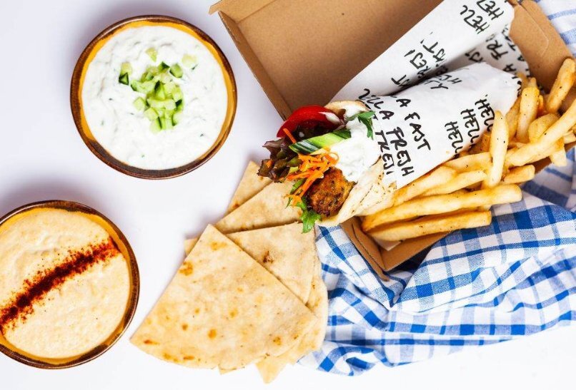 A souvlaki and chips in a box on top of a blue and white chequered cloth. To the left is a bowl of tzatziki, a bowl of rice pudding and a pile of pita bread triangles.